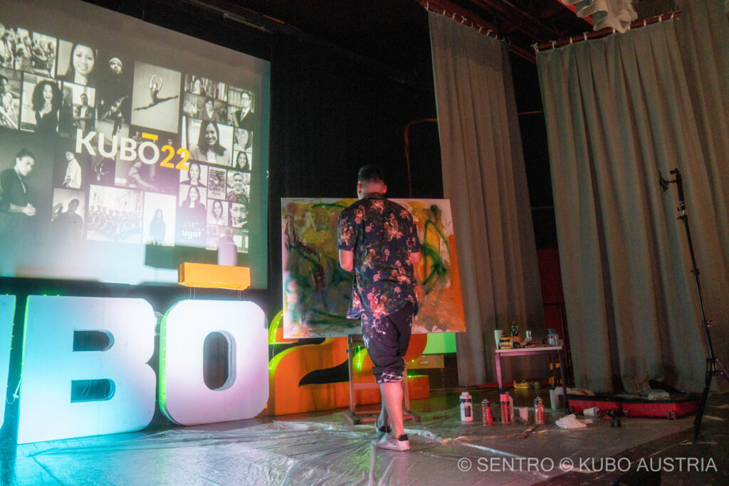 Ein junger Künstler steht auf einer Bühne vor einer Staffelei und malt ein Bild, während er sich von groovige Musik inspirieren lässt.